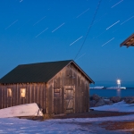 Fish House Under Stars: The lights remain on at night as workers toil to clean fish in a traditional cedar, fish house in the Grand Marais, Minnesota harbor. The lighthouse calls out to boats sailing Lake Superior. The Coast Guard building remains lit up under the startrails.
