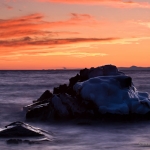 The Rock and the Shore: At sunset waves wash around an ice-covered rock. The Sawtooth Mountains are in the distance. Lake Superior, Minnesota.