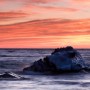 An ice-coated rock awash with waves under a reddish-orange sky.