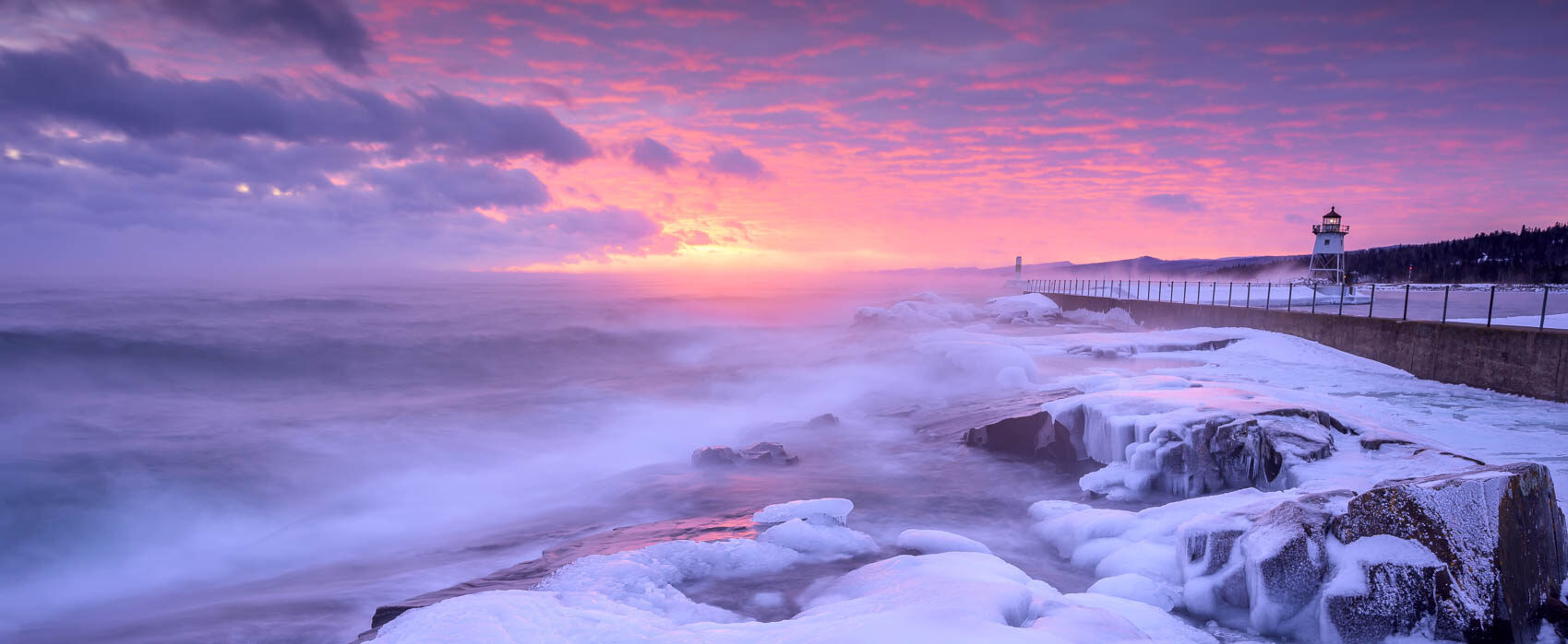 An icy shoreline painted pink at sunset. The Grand Marais lighthouse is in the distance.