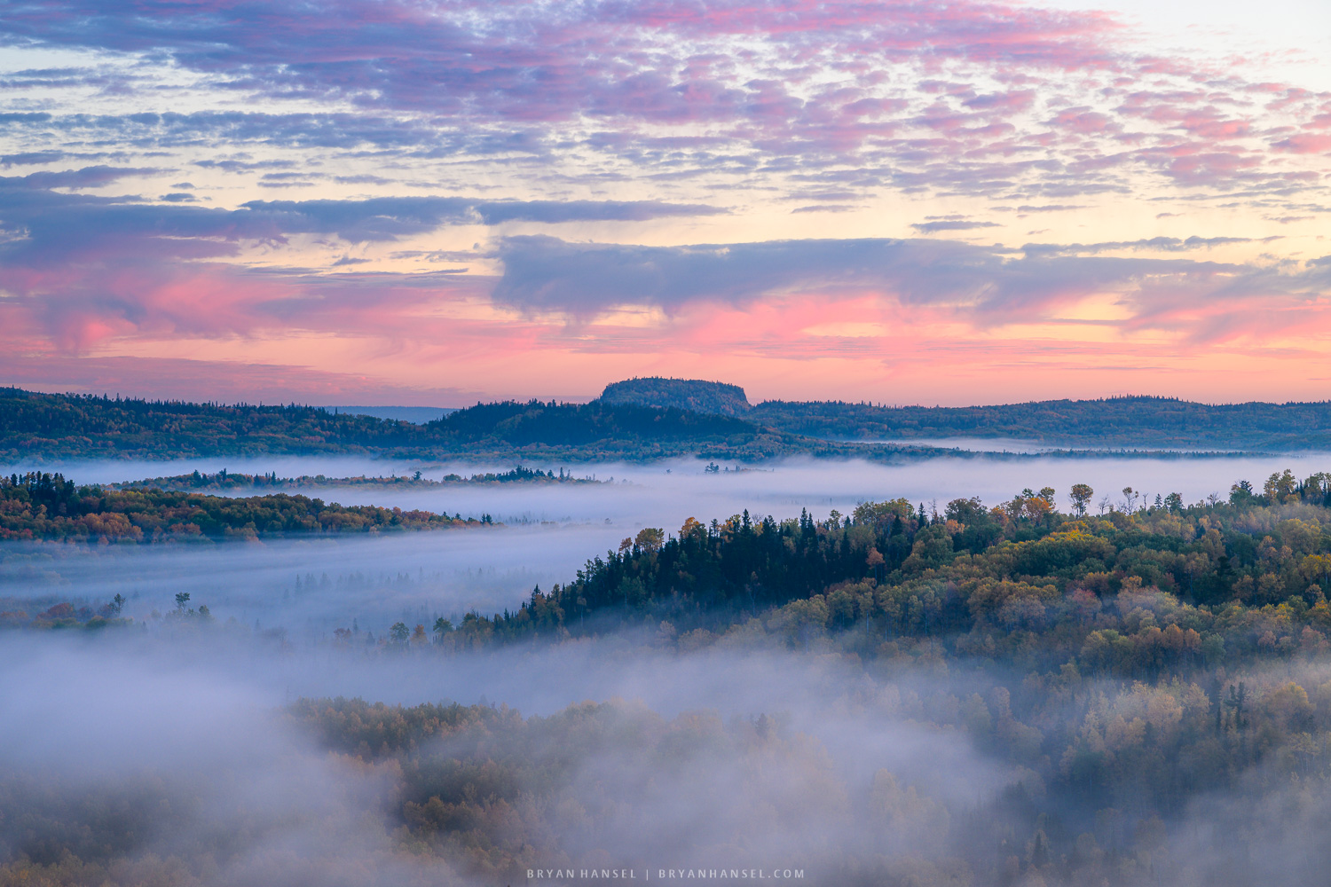 A foggy valley with hills and buttes coming out of the fog under the sunrise clouds.